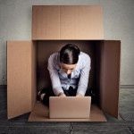 Closeup young businesswoman hiding sitting in the carton box working on laptop computer